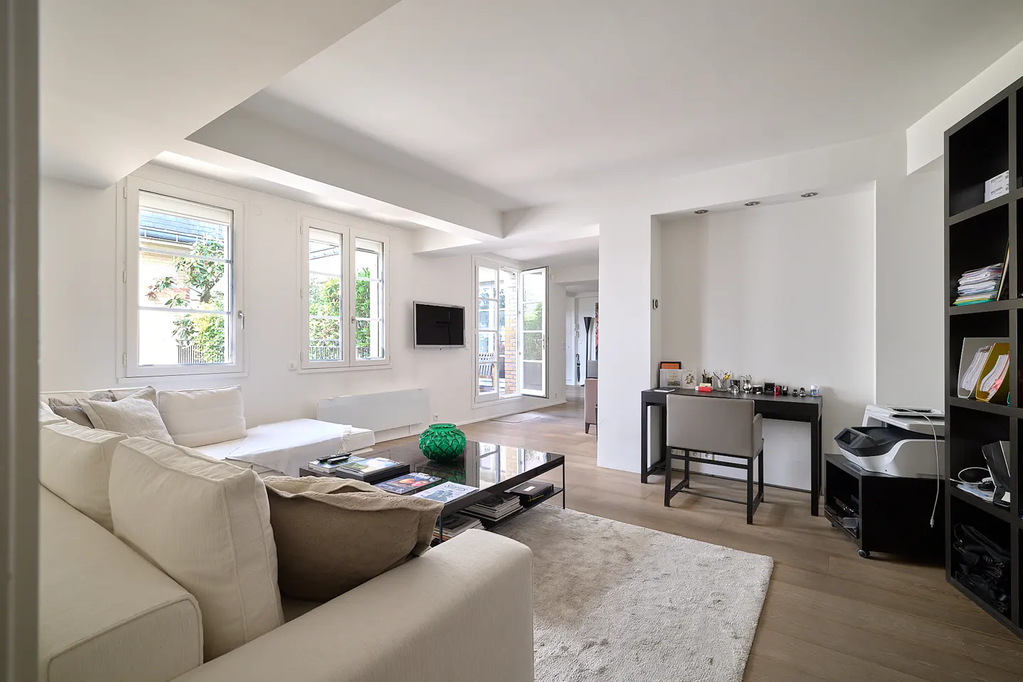 Bright living room with white walls, wood floors, and large windows. A white sofa faces a black coffee table with a green vase. A desk and bookcase are on the right.