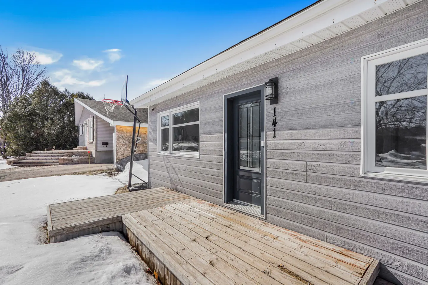 Exterior view of a gray house with a wooden deck, black door, and white windows under a blue sky. A basketball hoop is visible in the background.