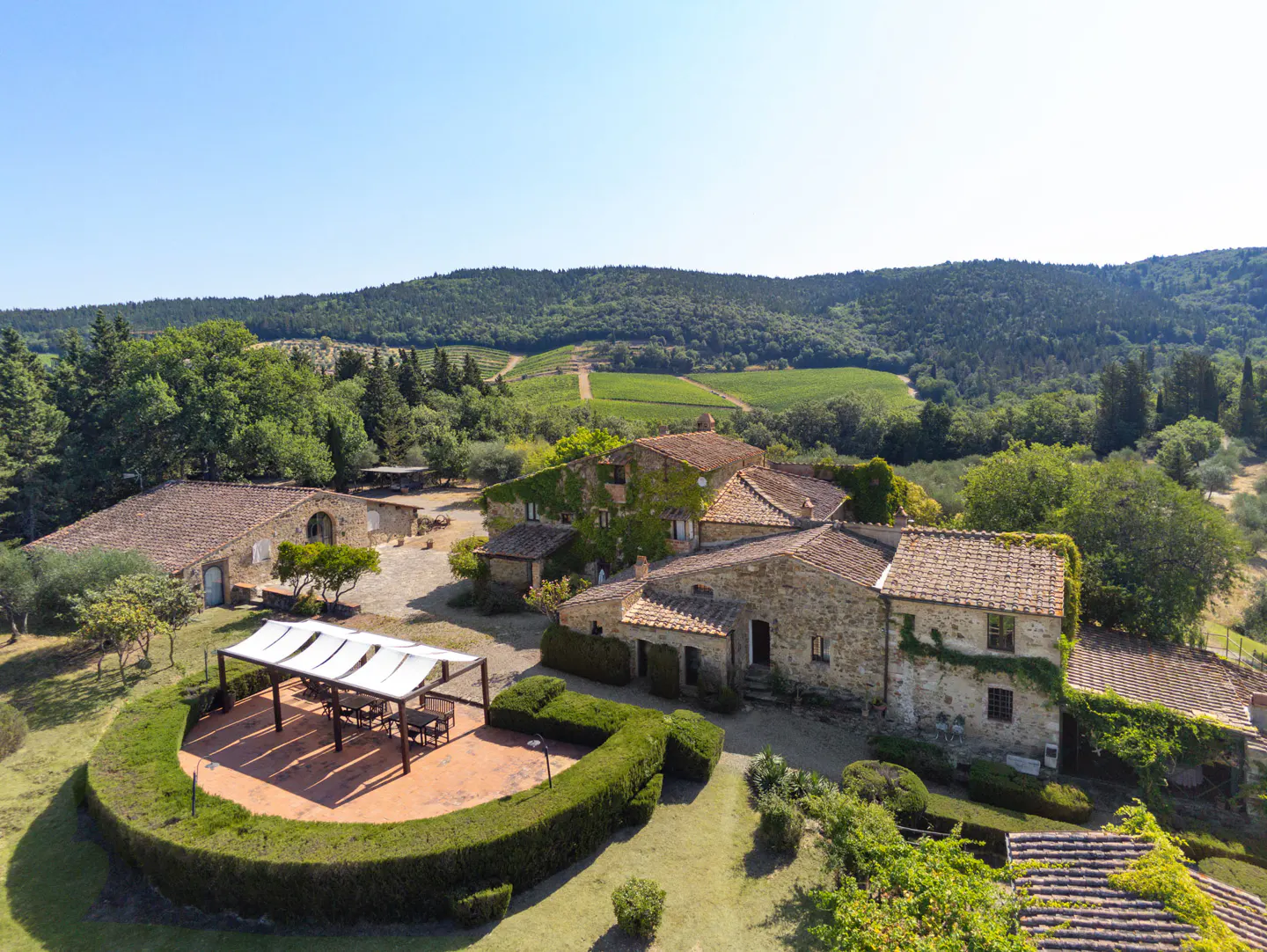 Aerial view of a Tuscan villa with stone buildings, red tile roofs, and a pergola-covered patio surrounded by green lawns and trees. Rolling hills in the background.