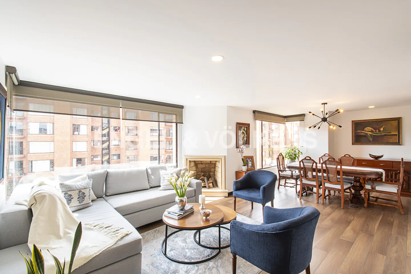 Bright, open-concept living and dining area with gray sofa, blue chairs, and wood dining table. Large windows offer city views.