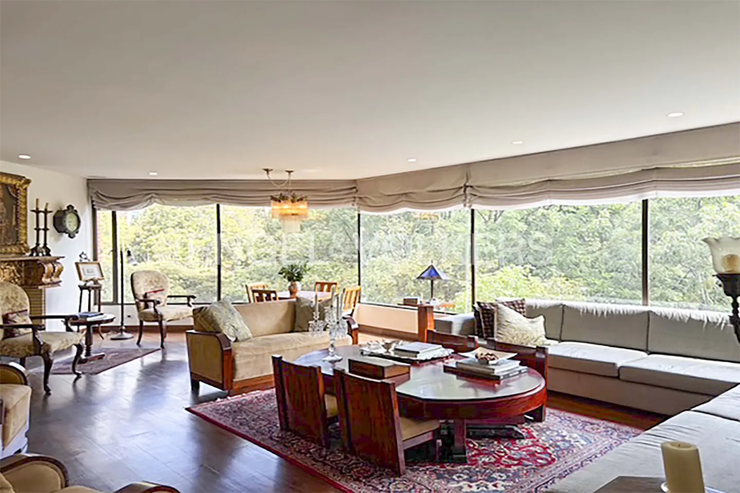Spacious living room with a large oval wooden table, beige sofas, and floor-to-ceiling windows overlooking lush greenery.