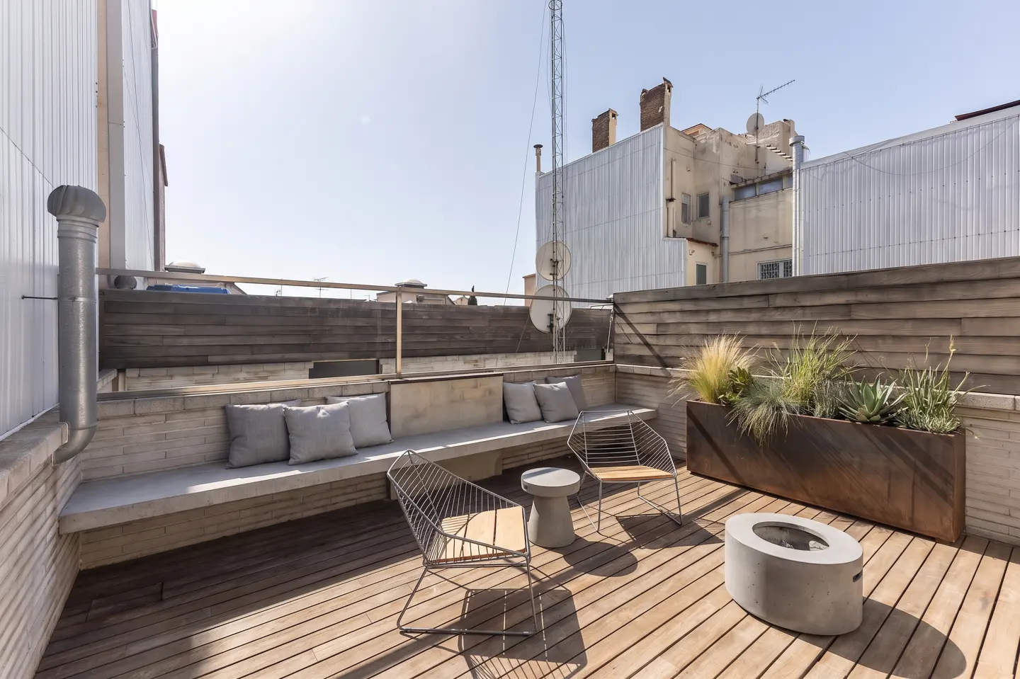 Rooftop patio with wooden deck, built-in seating with gray cushions, metal chairs, and a planter box with greenery. Buildings in the background.