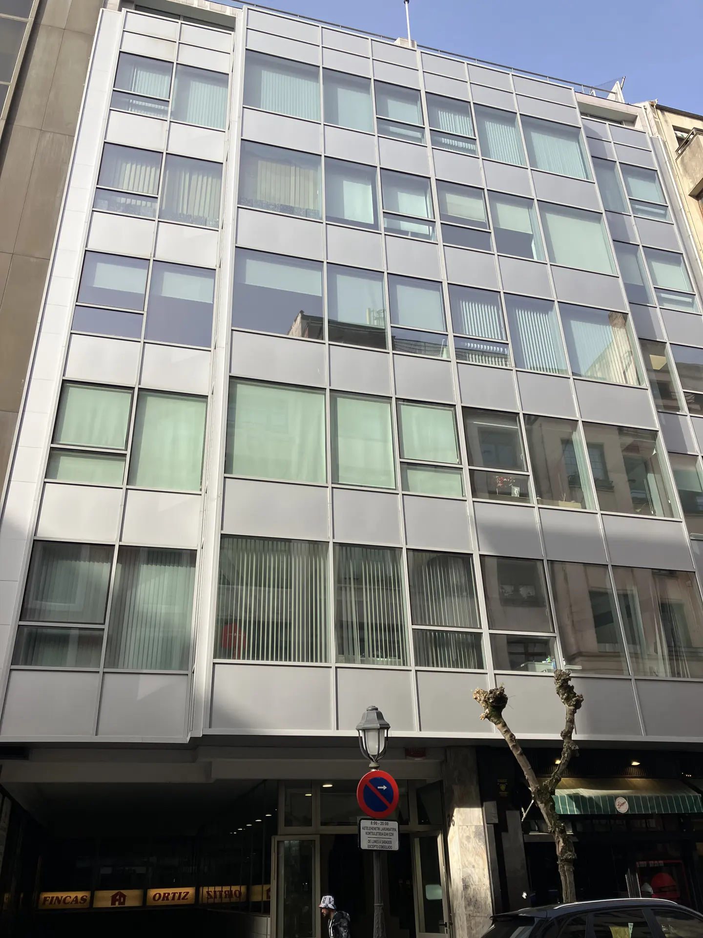 Exterior of a modern, multi-story office building with a gray facade and large windows. Street signs and a tree are visible in the foreground.