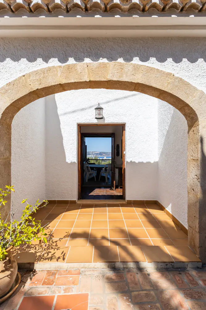 Stone archway frames a view of the sea from a tiled patio. White walls and a terracotta tiled floor create a bright, inviting space.