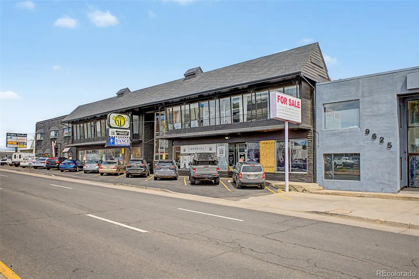 A two-story commercial building with a "For Sale" sign, parked cars, and a gray stucco building next door under a blue sky.