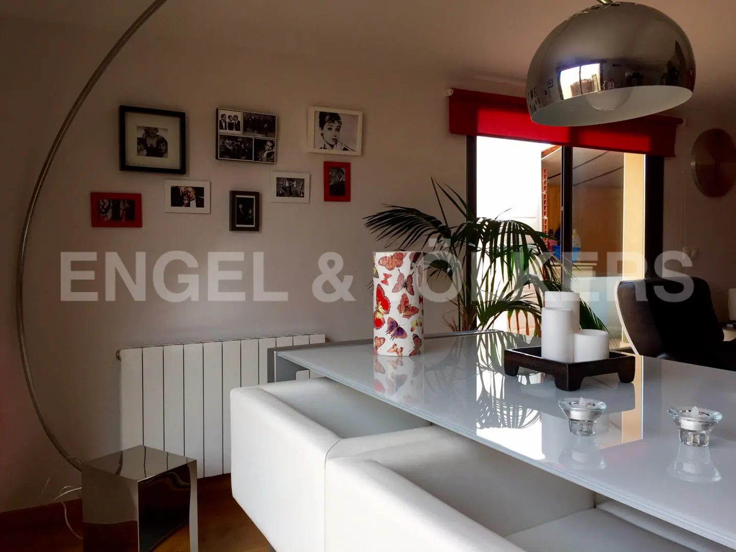Interior view of a dining area with a white table, bench seating, and a silver arc lamp. Family photos adorn the wall. A plant and candles decorate the table.