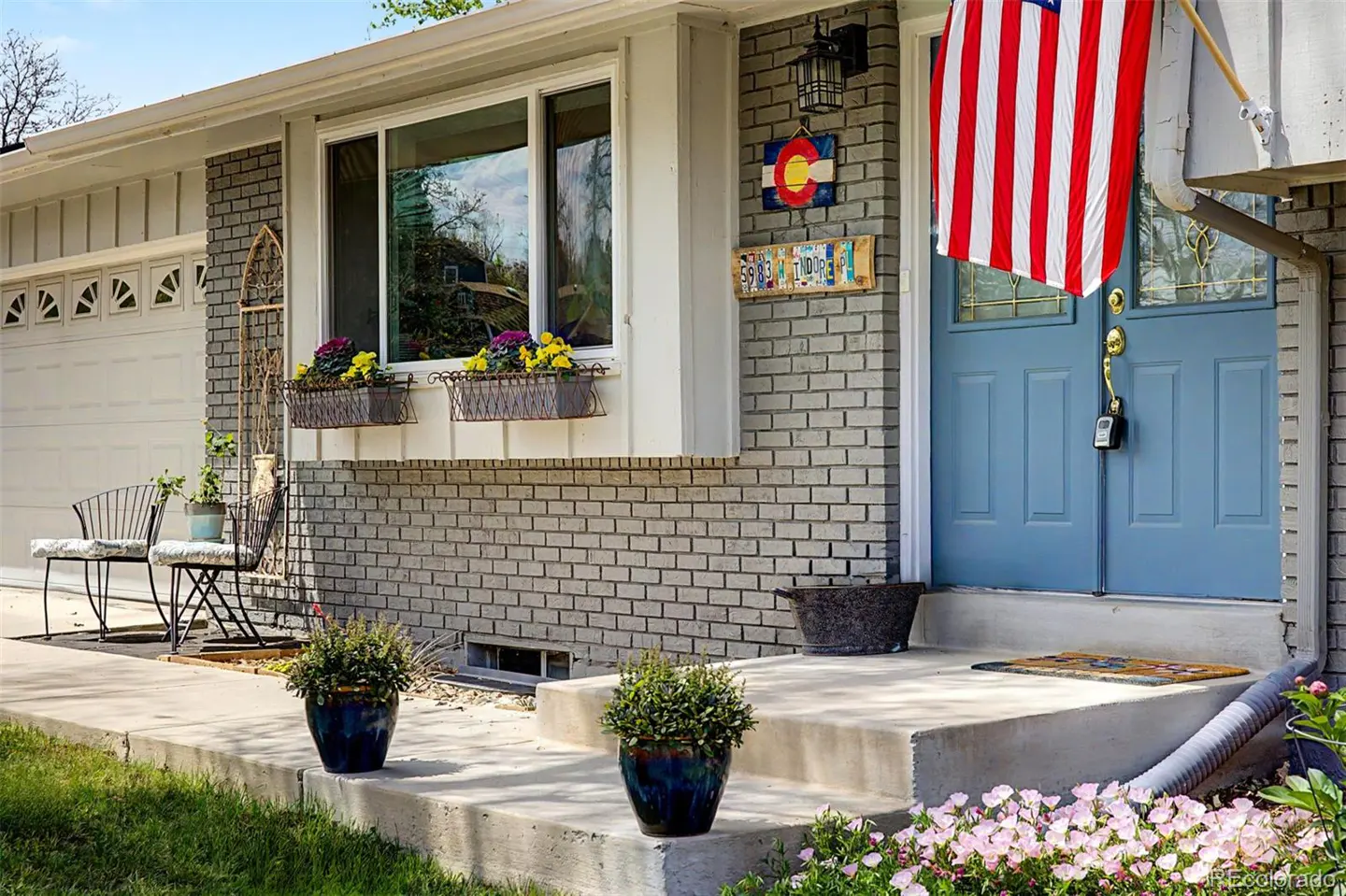 Exterior of a gray brick house with a blue door and American flag. Flower boxes sit under a window. Two blue pots sit on the concrete porch.