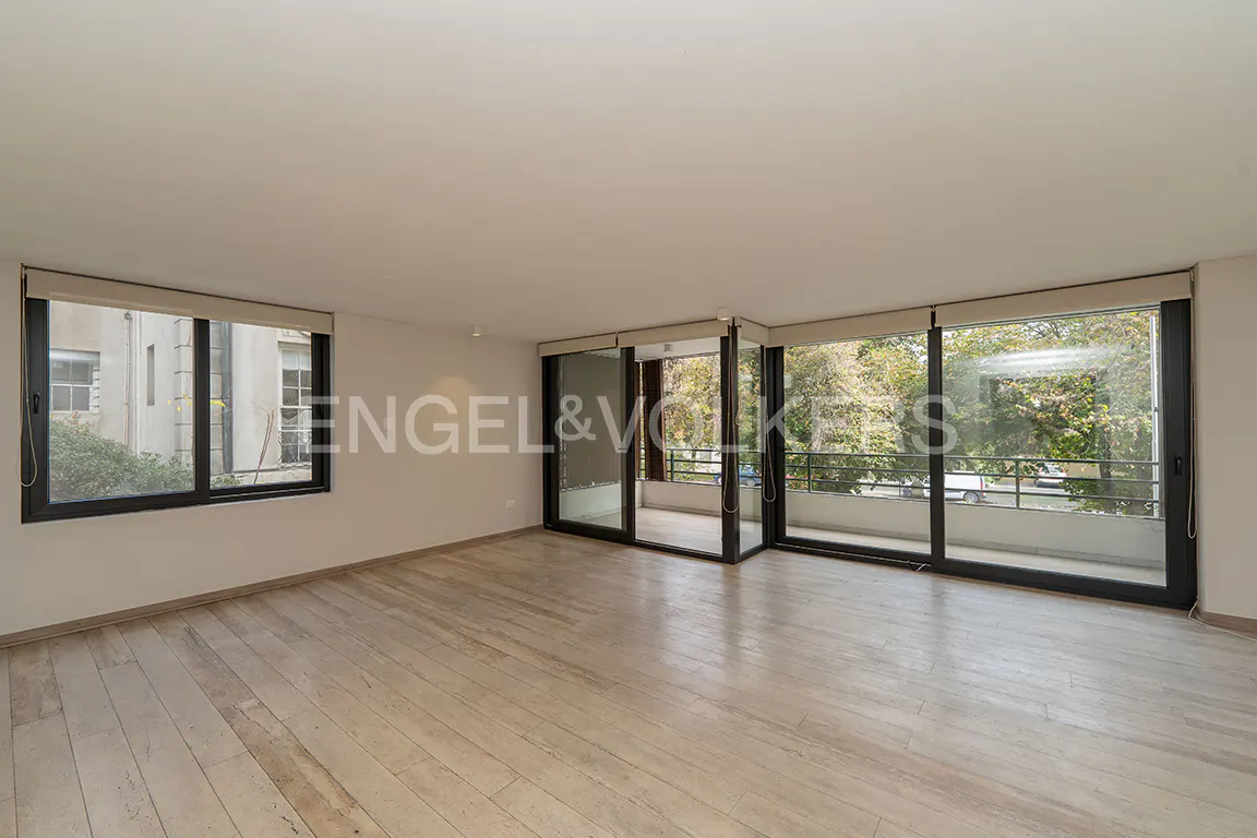 Empty living room with light wood floors, white walls, and large windows overlooking a balcony and trees. Engel & Volkers logo visible.