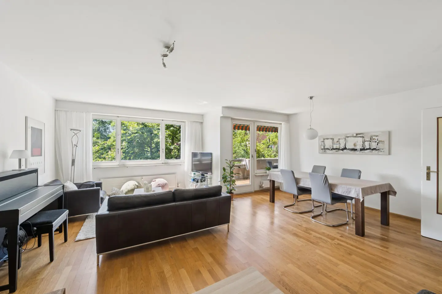 Bright, open-plan living and dining area with wood floors, piano, and balcony. Brown sofa faces large windows with green trees outside.