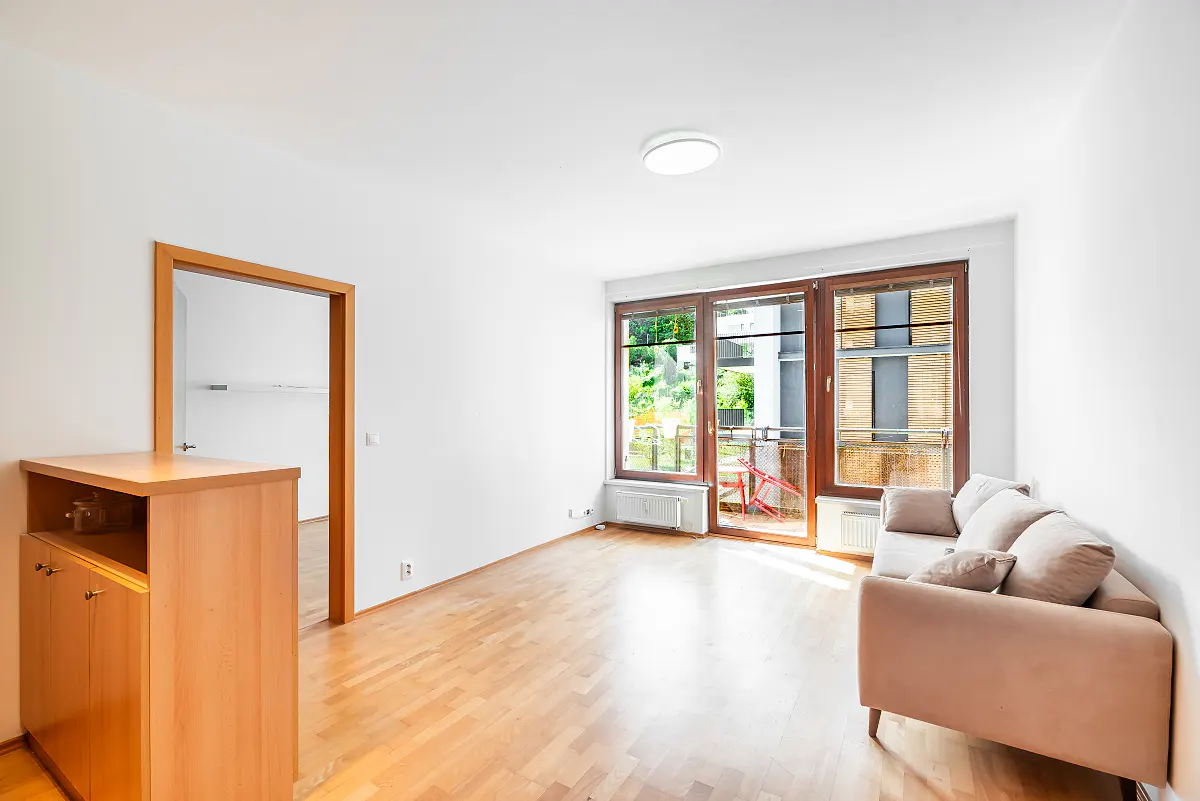Bright living room with wood floors, white walls, and a beige sofa. A door leads to another room, and a balcony is visible through large windows.