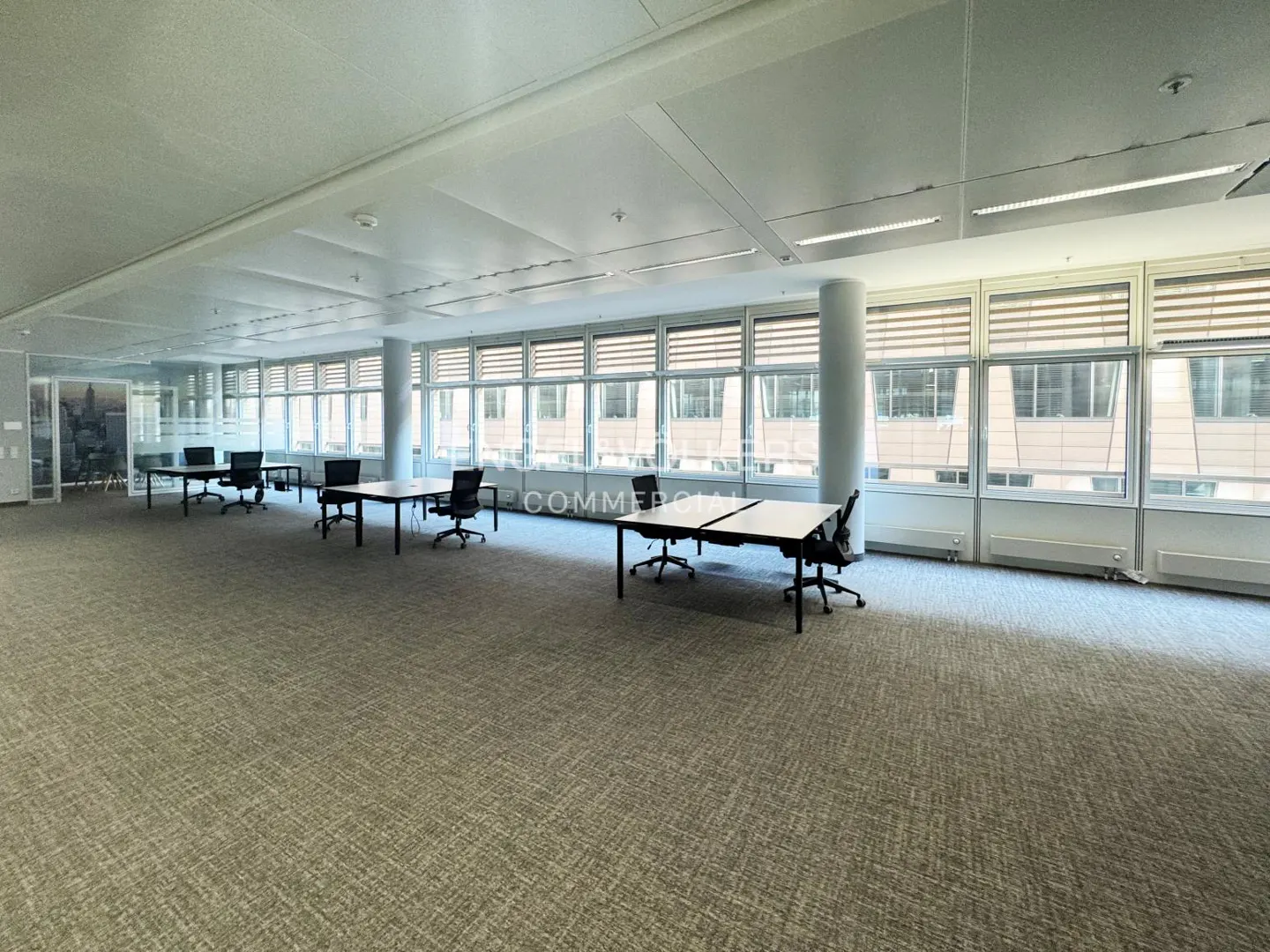 Bright, empty office space with gray carpet, black desks and chairs, and large windows with blinds.