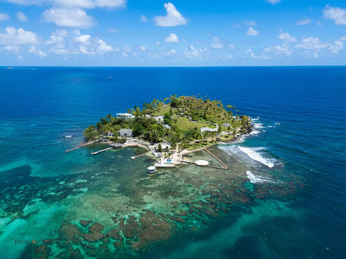 Aerial view of a tropical island with white buildings, palm trees, and turquoise water under a blue sky.