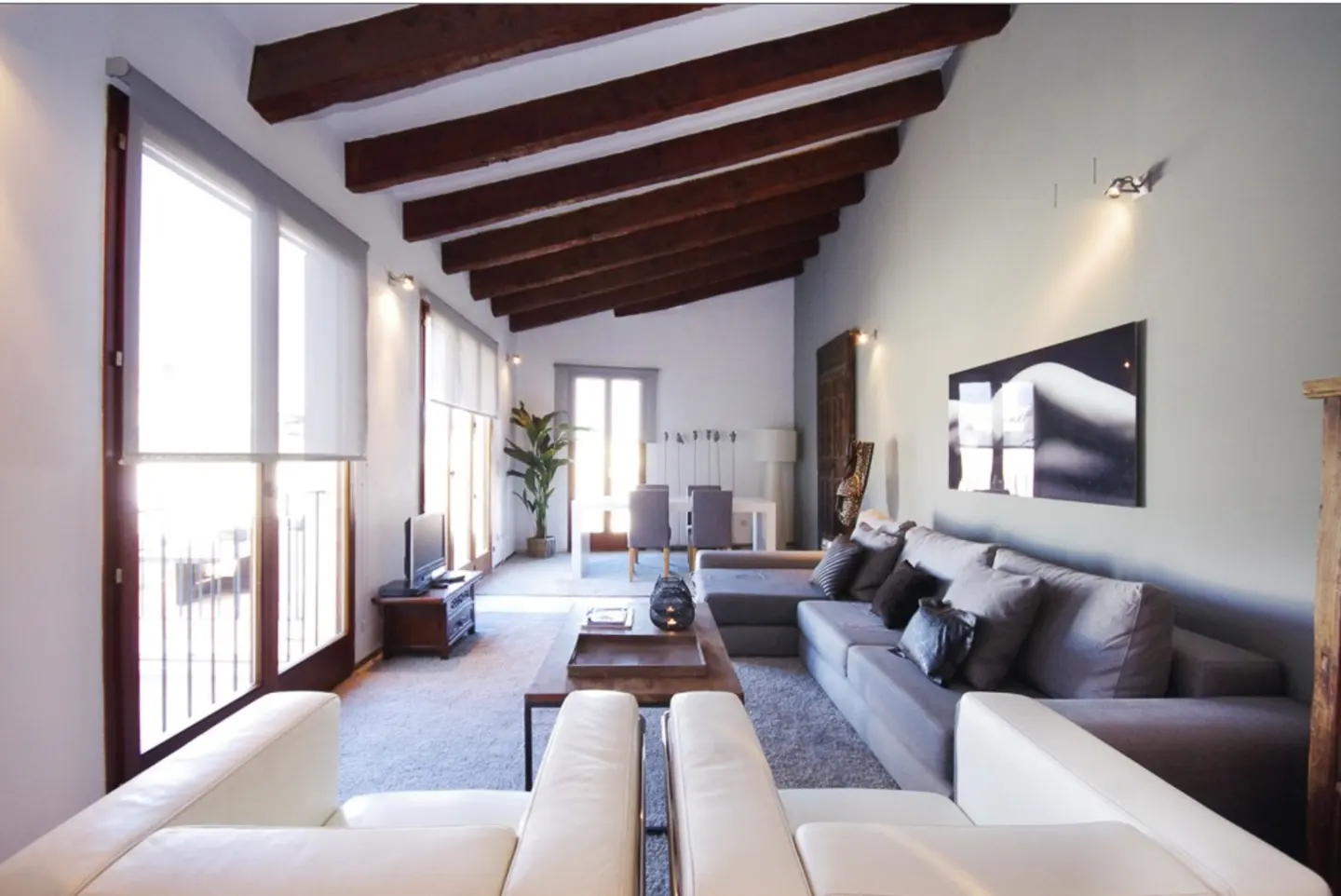 Modern living room with vaulted wood-beam ceiling, gray sectional sofa, white armchairs, and a dining area in the back.