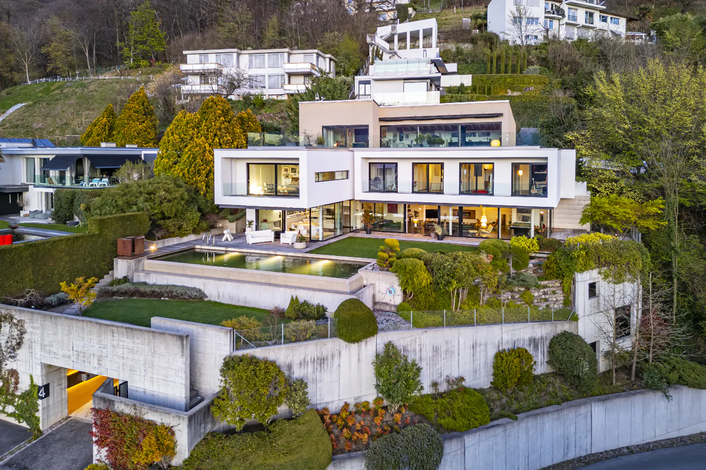 A modern white house with large windows sits on a green hillside with a pool and manicured gardens.