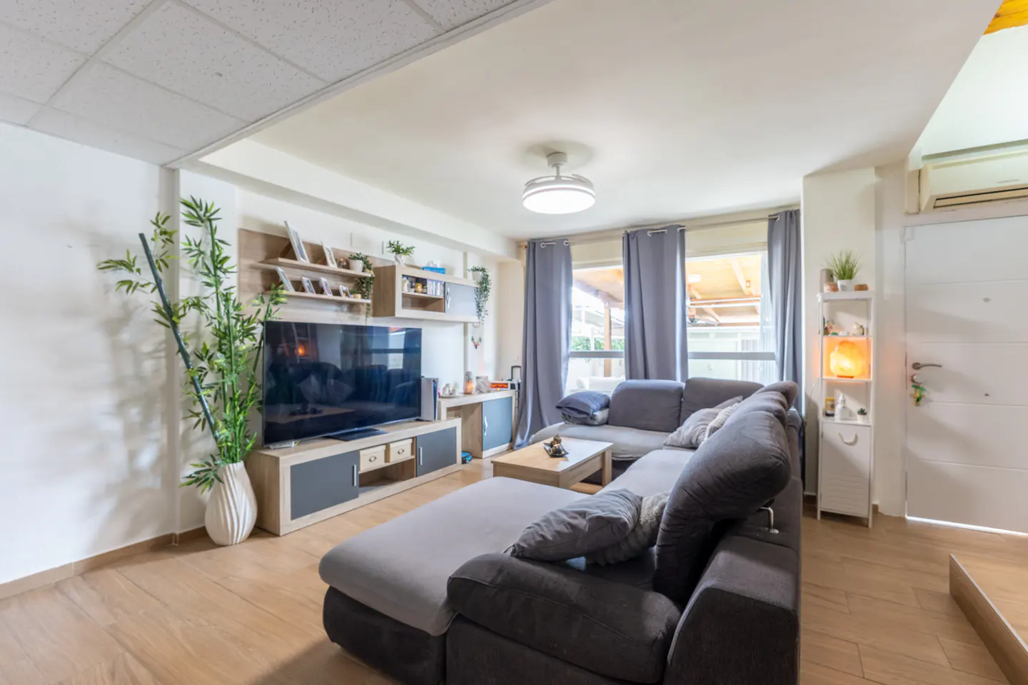 Living room with a gray sectional sofa, wood floors, TV, and shelves. Gray curtains frame the window.