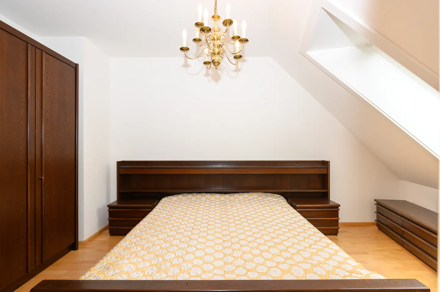 Bedroom with a wooden bed, yellow patterned bedding, and matching furniture. A gold chandelier hangs from the white ceiling.