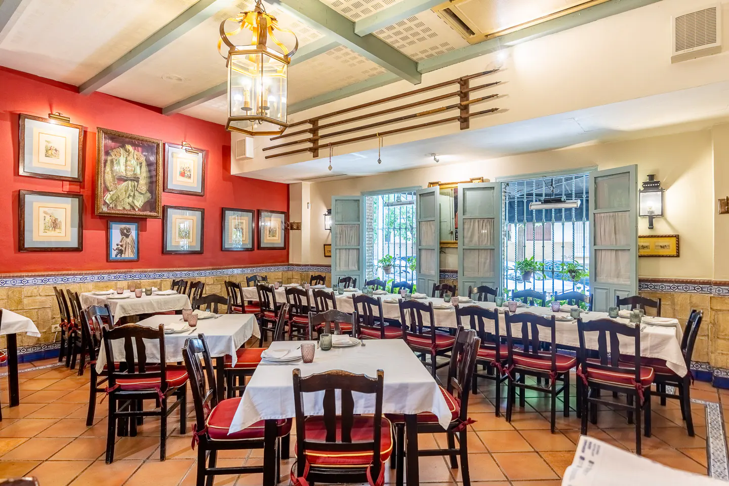A restaurant interior with red walls, framed artwork, wooden chairs with red cushions, and tables set with white tablecloths.