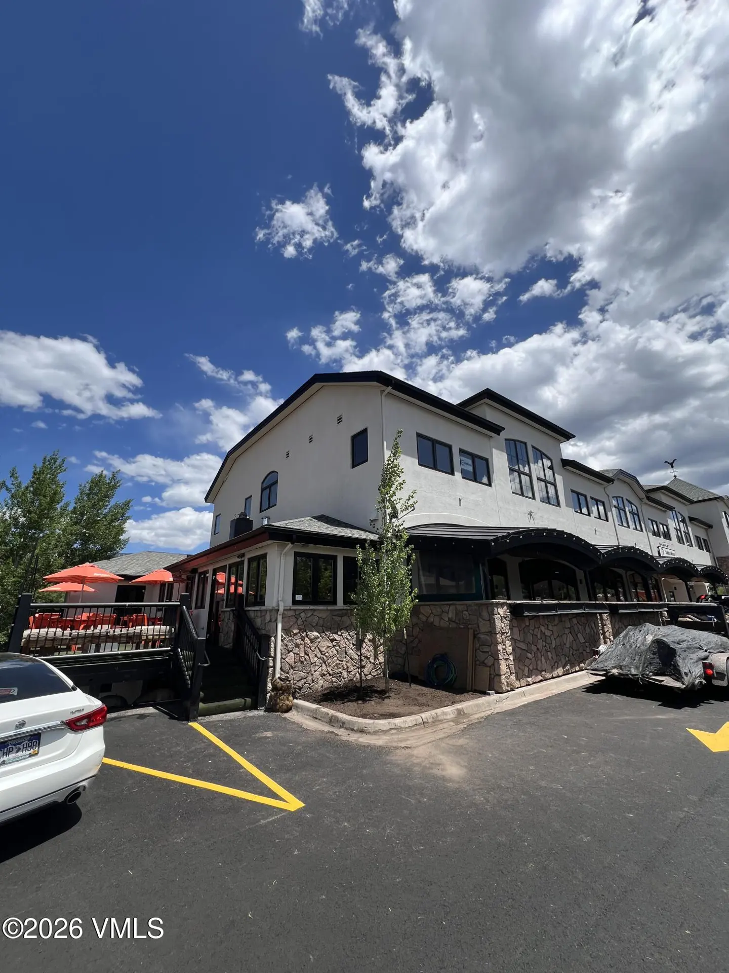 Exterior view of a white commercial building with stone accents, a patio with orange umbrellas, and a parking lot.