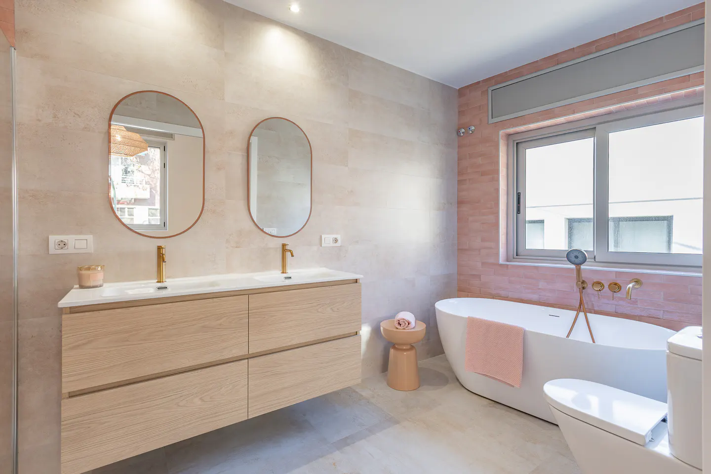 Modern bathroom with double sink vanity, oval mirrors, and a freestanding white tub by a window with pink brick accent wall.