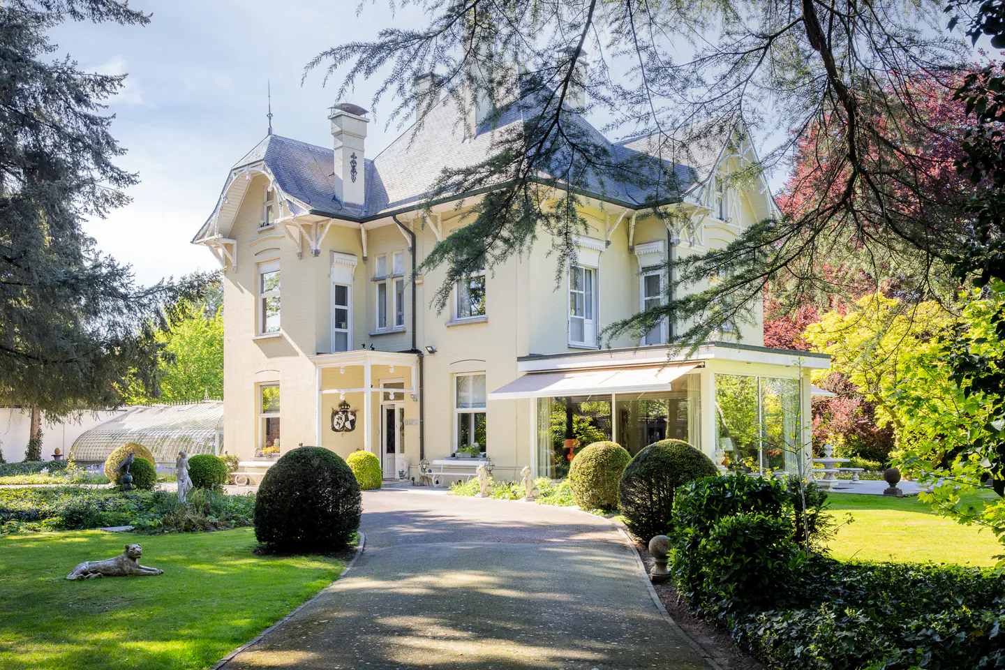 A large, pale yellow house with a gray roof, surrounded by green lawns and trees. A stone driveway leads to the entrance.