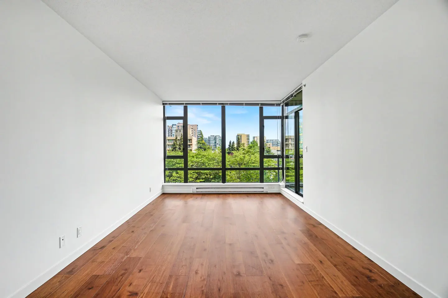 Bright, empty room with hardwood floors and white walls. Large window shows city buildings and green trees.