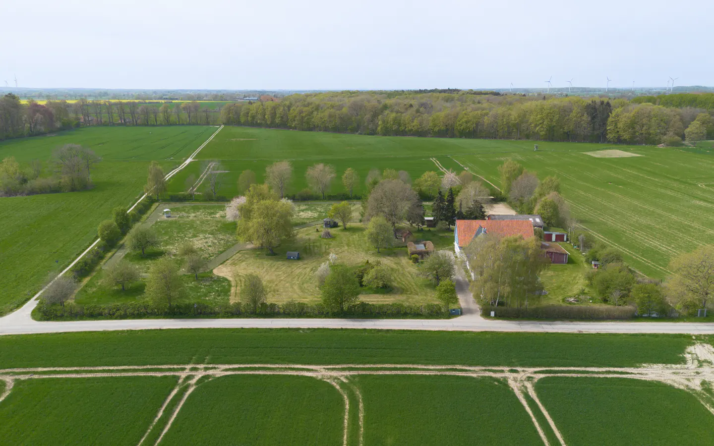 Aerial view of a rural property with a red-roofed house, surrounded by green fields and trees. A road runs in front of the property.