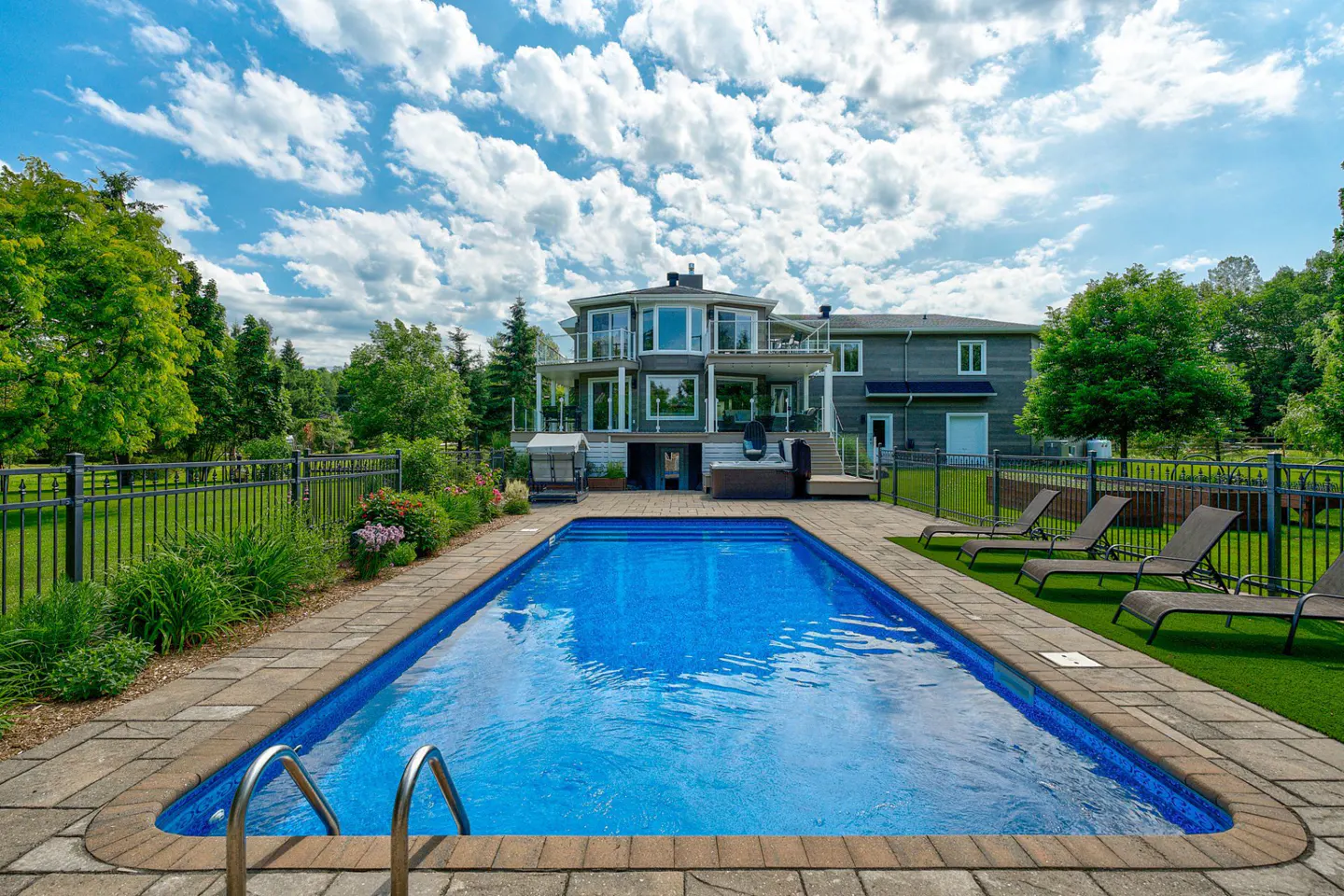 A rectangular blue swimming pool is centered in a backyard with lounge chairs and a gray two-story house.
