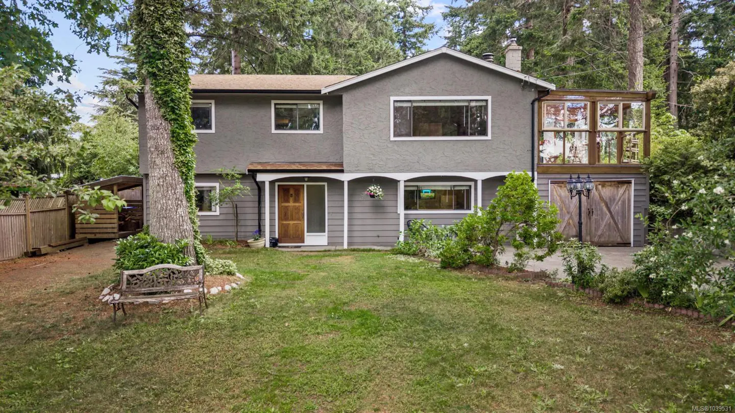 Two-story grey house with a wooden front door, a sunroom, and a metal bench in a grassy yard surrounded by tall trees.
