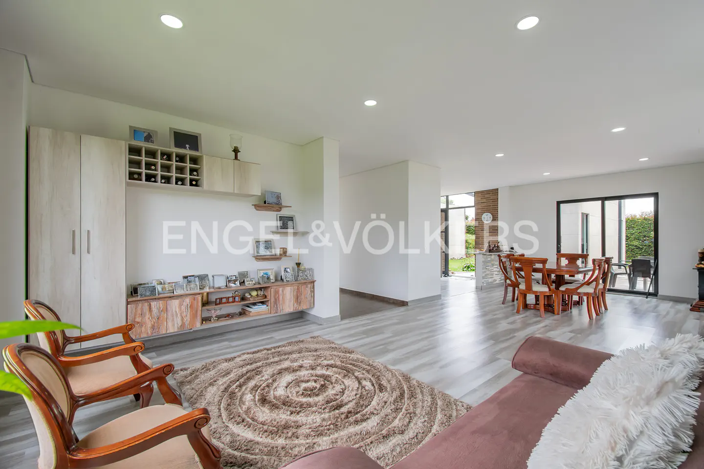 Bright living room with wood floors, brown sofa, and chairs. A large rug sits in the center, with built-in shelves and cabinets on the wall.