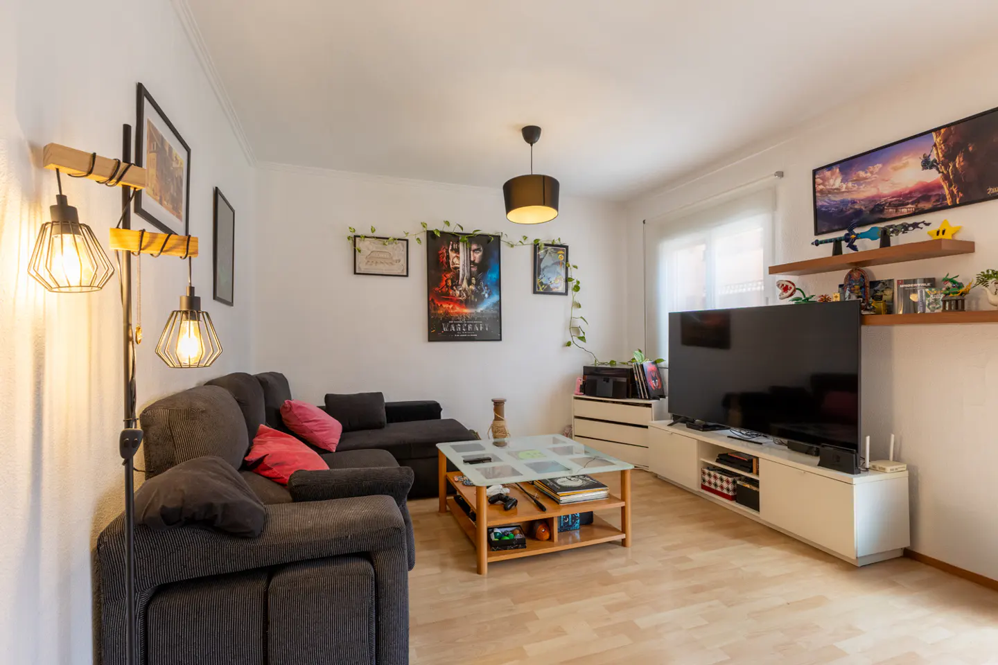 Living room with a gray sectional sofa, wood floors, and a large TV. Posters and shelves decorate the white walls.