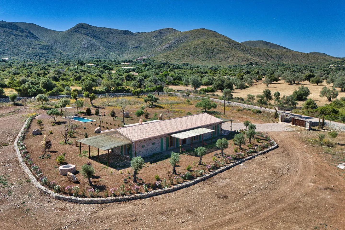 Aerial view of a stone house with a red tile roof, pool, and mountain backdrop under a clear blue sky.