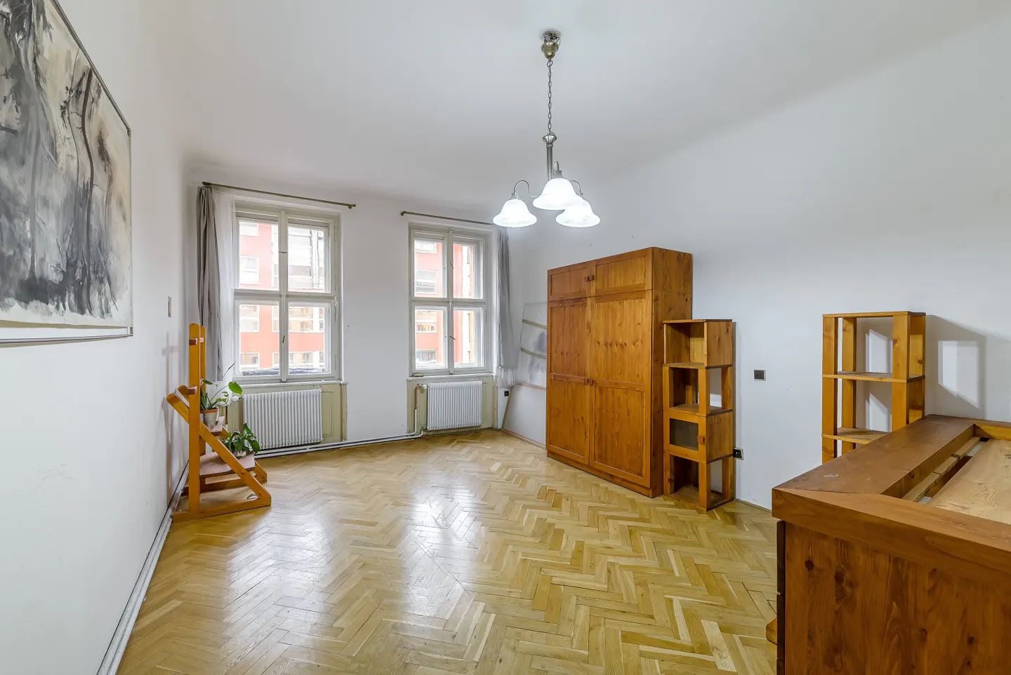 Bright room with herringbone wood floors, white walls, and two windows. A wooden wardrobe and shelves stand against the wall. A modern chandelier hangs from the ceiling.