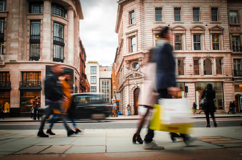 Busy London street scene with blurred pedestrians and a black taxi. Buildings are light stone with many windows.