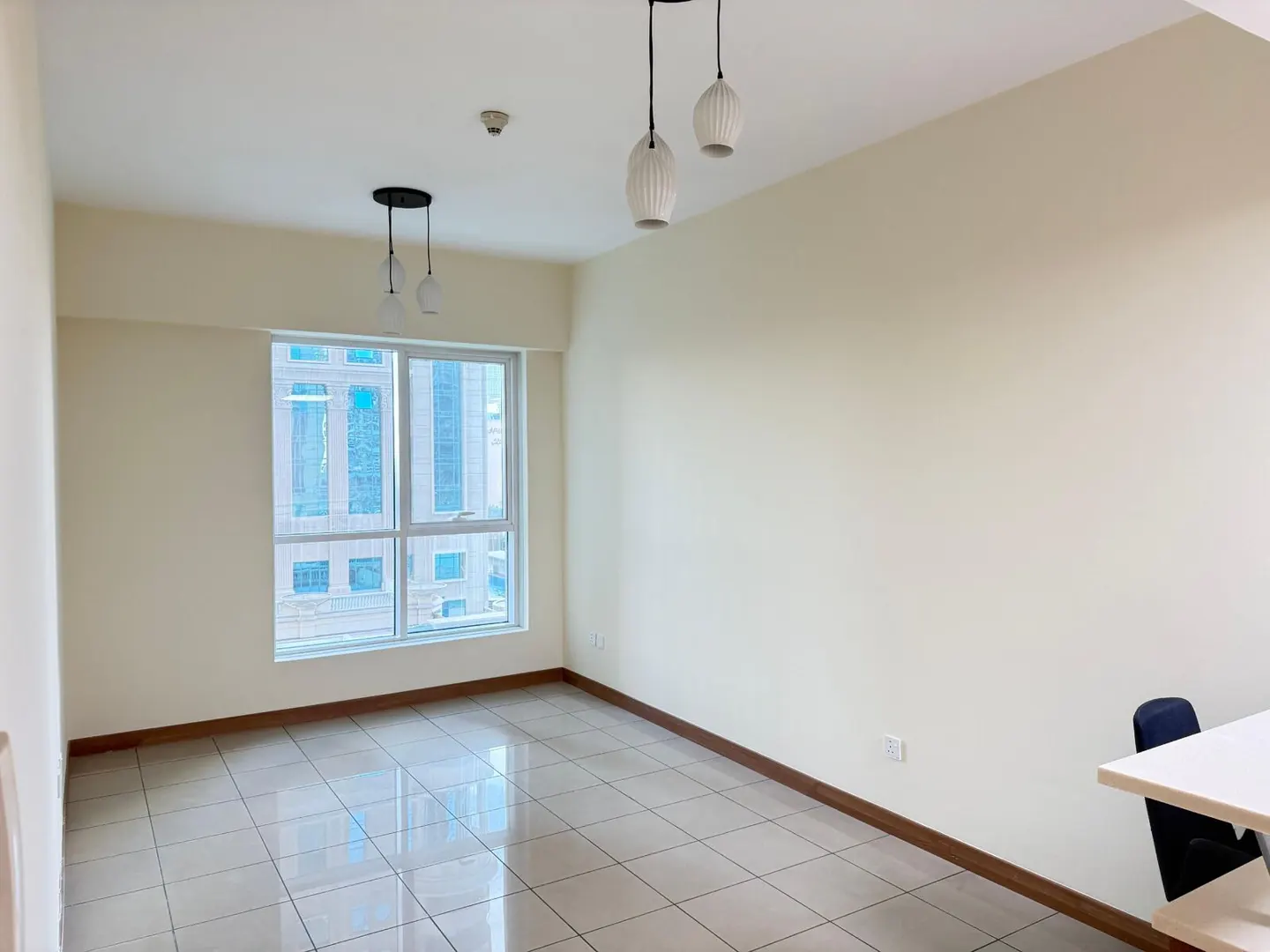 Empty room with beige walls, tiled floor, and a window showing city buildings. Two pendant lights hang from the ceiling.
