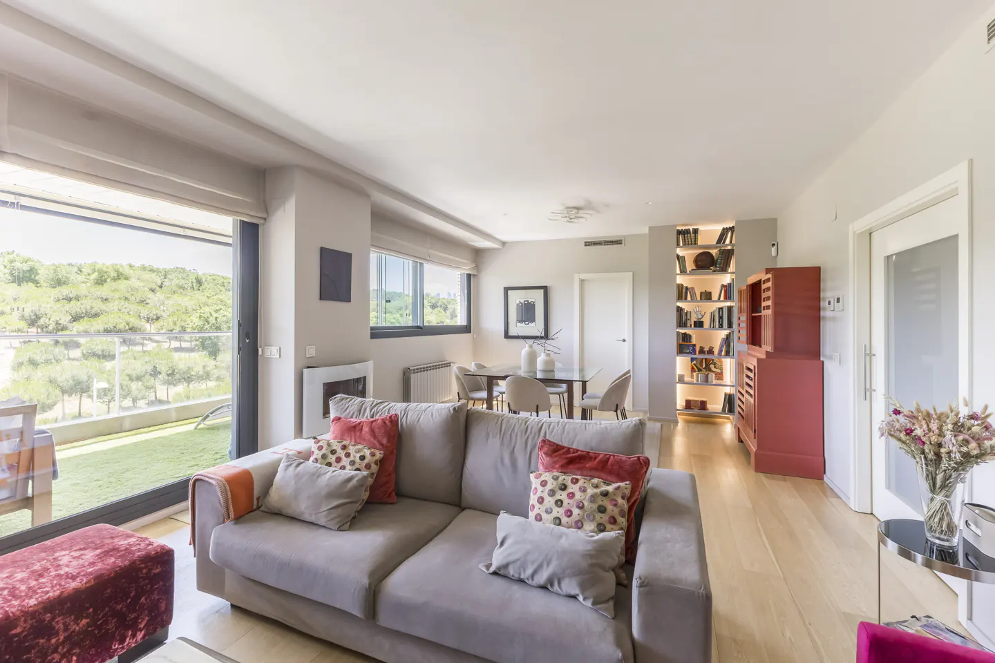 Bright living room with a grey sofa, red accents, a dining area, and a large window overlooking a green landscape.