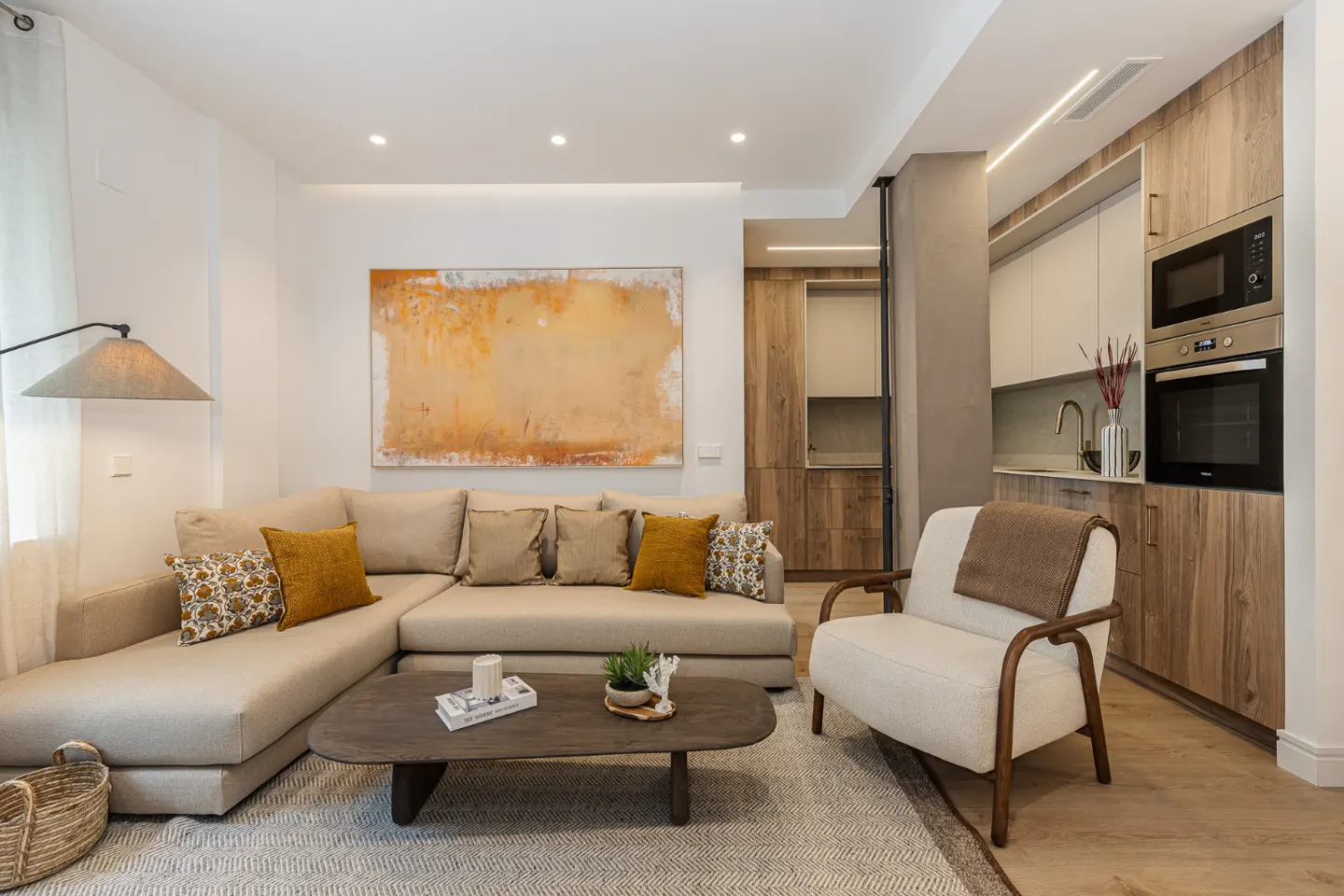 Bright living room with beige sectional, wood coffee table, and white armchair near a modern kitchen. An abstract painting hangs on the wall.