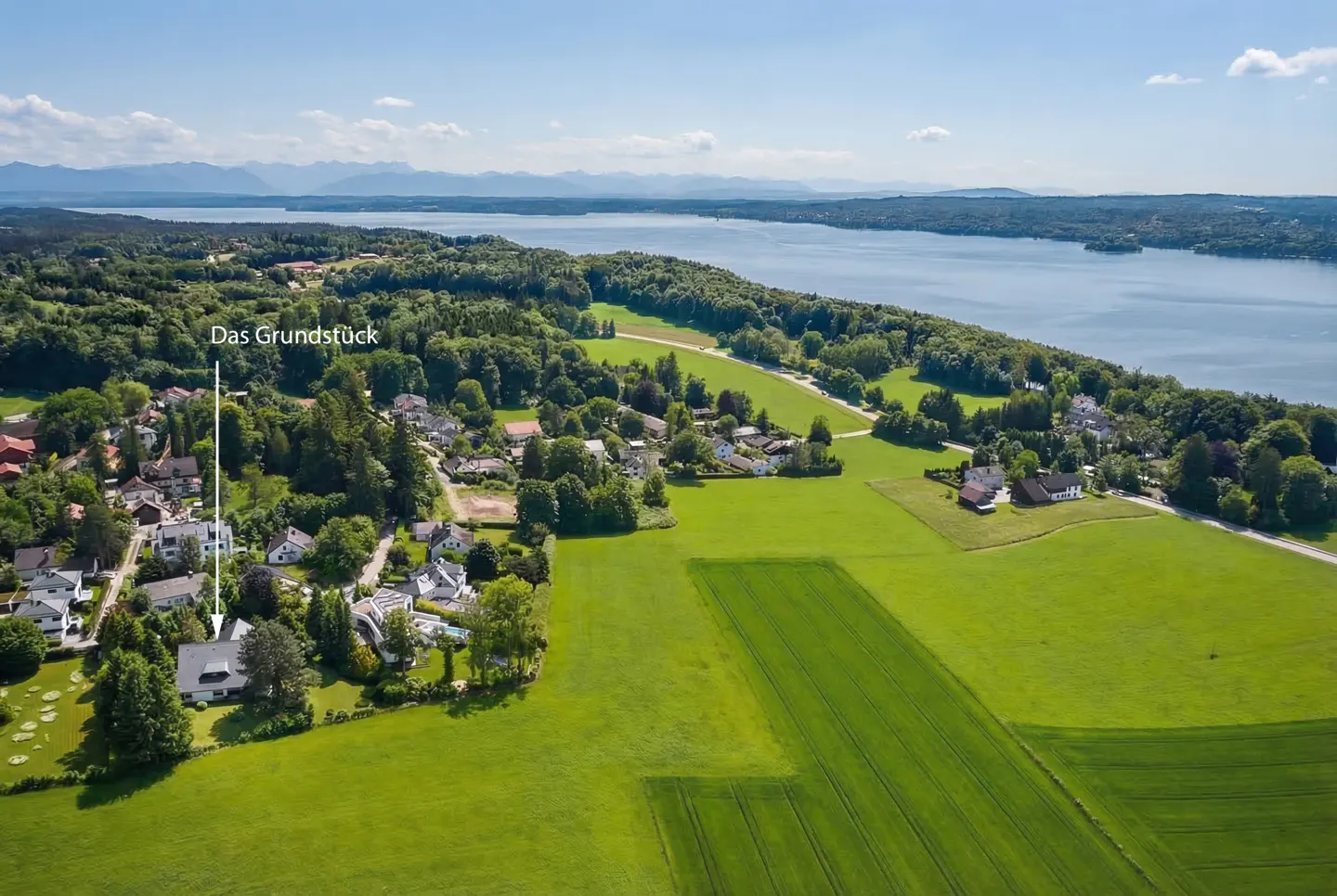 Aerial view of a property, "Das Grundstück," near a lake, surrounded by green fields and trees under a blue sky.