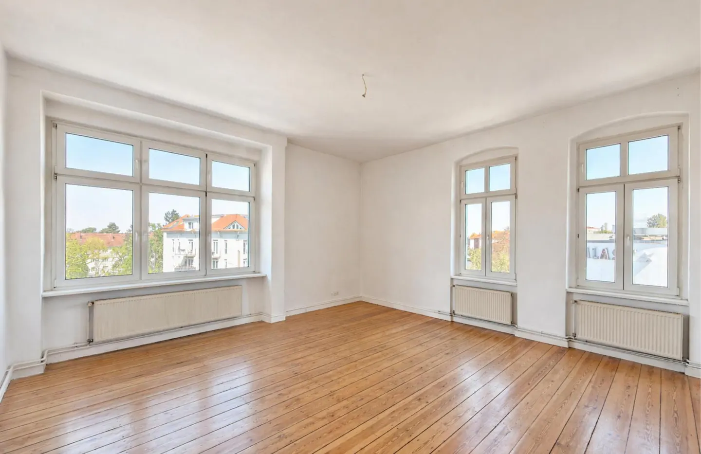Bright, empty room with hardwood floors, white walls, and three large windows showing a neighborhood scene.