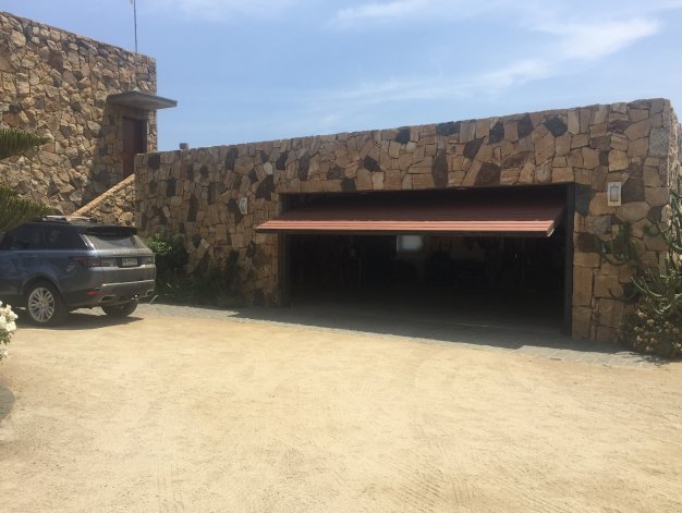 Stone house with a partially open garage door. A gray SUV is parked on the gravel driveway. Blue sky in the background.