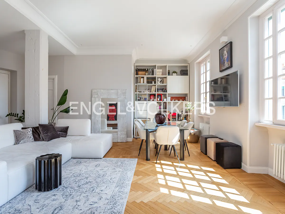 Bright living room with white sofa, dining table, and herringbone wood floors. Bookshelves and a TV are on the wall.