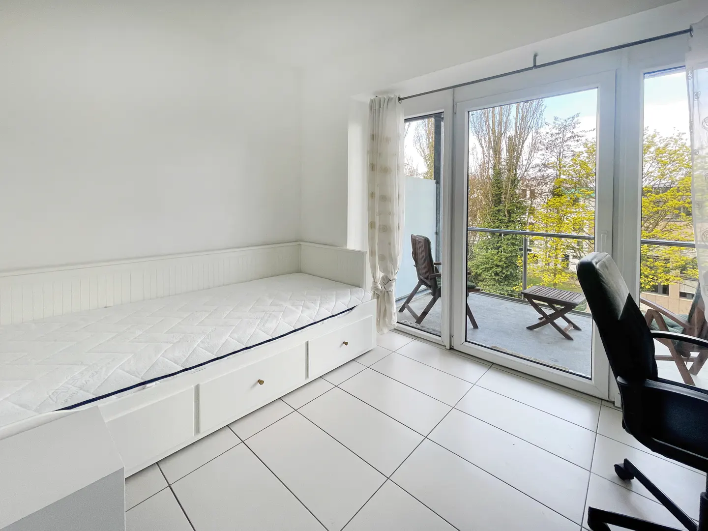 Bright room with white tile floor, daybed, and balcony. Balcony has chairs and a table, with trees visible beyond. A black office chair is in the room.