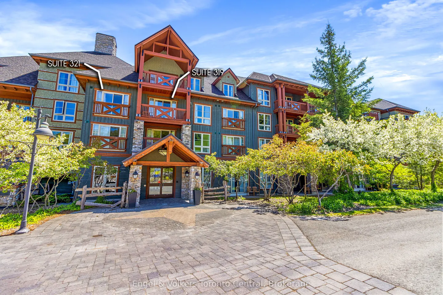 Exterior of a multi-story building with blue siding, wood trim, balconies, and a stone entrance. Trees and a paved driveway are in front.