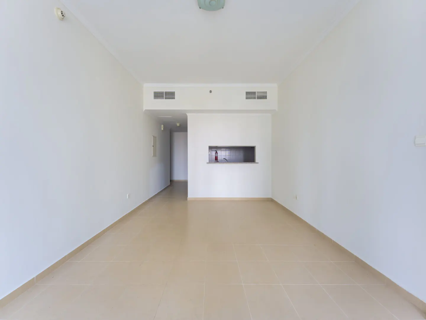 An empty, minimalist room with white walls, beige tiled floors, and a small kitchen pass-through window.