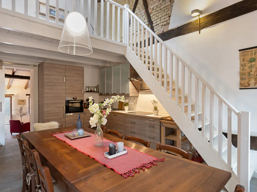 Interior view of a dining room with a long wooden table, chairs, and a red runner. A white staircase leads to a loft above the kitchen area.