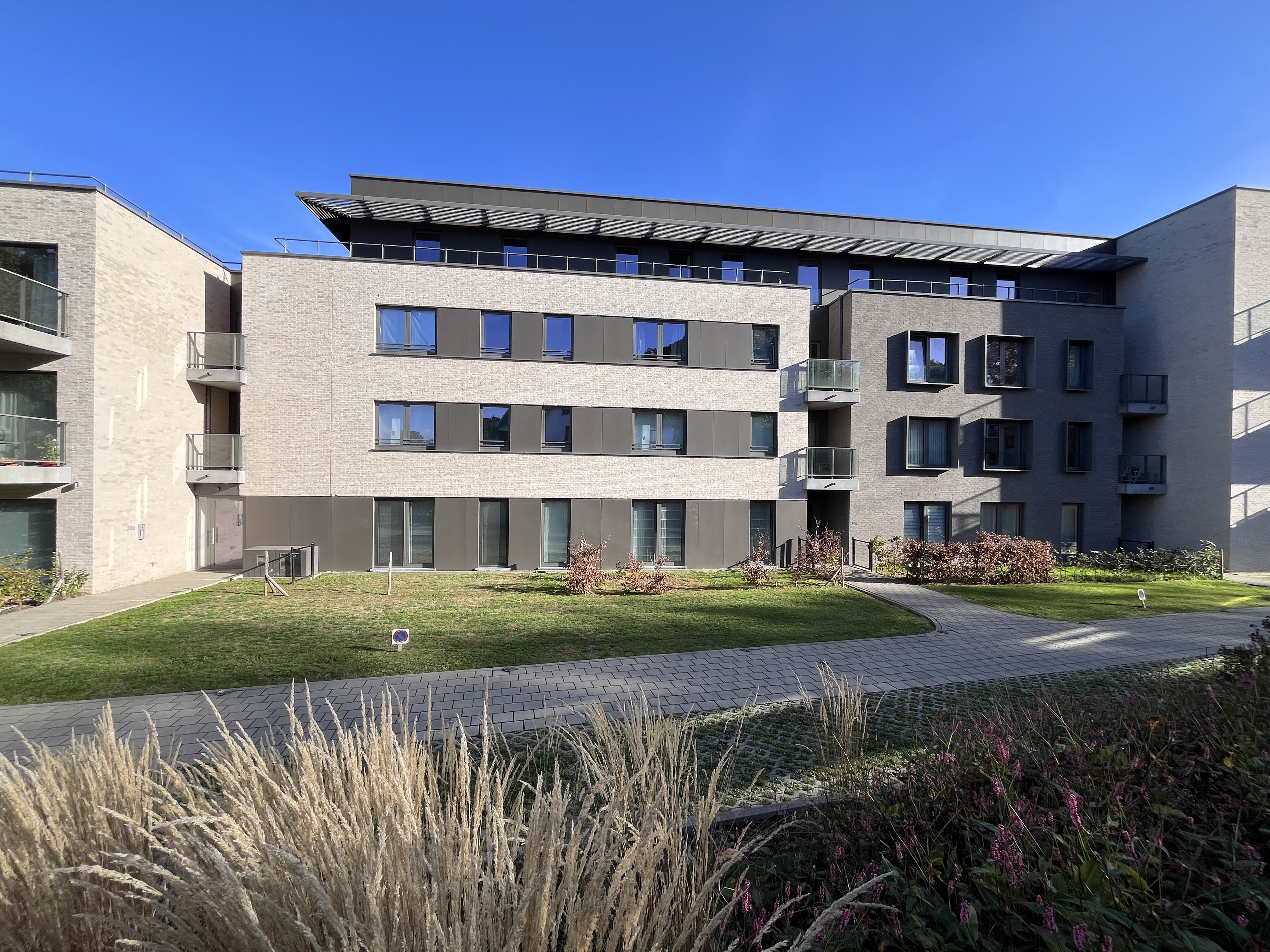 Modern apartment building with light brick and dark grey accents, featuring balconies and a landscaped lawn under a blue sky. [AI-generated text]