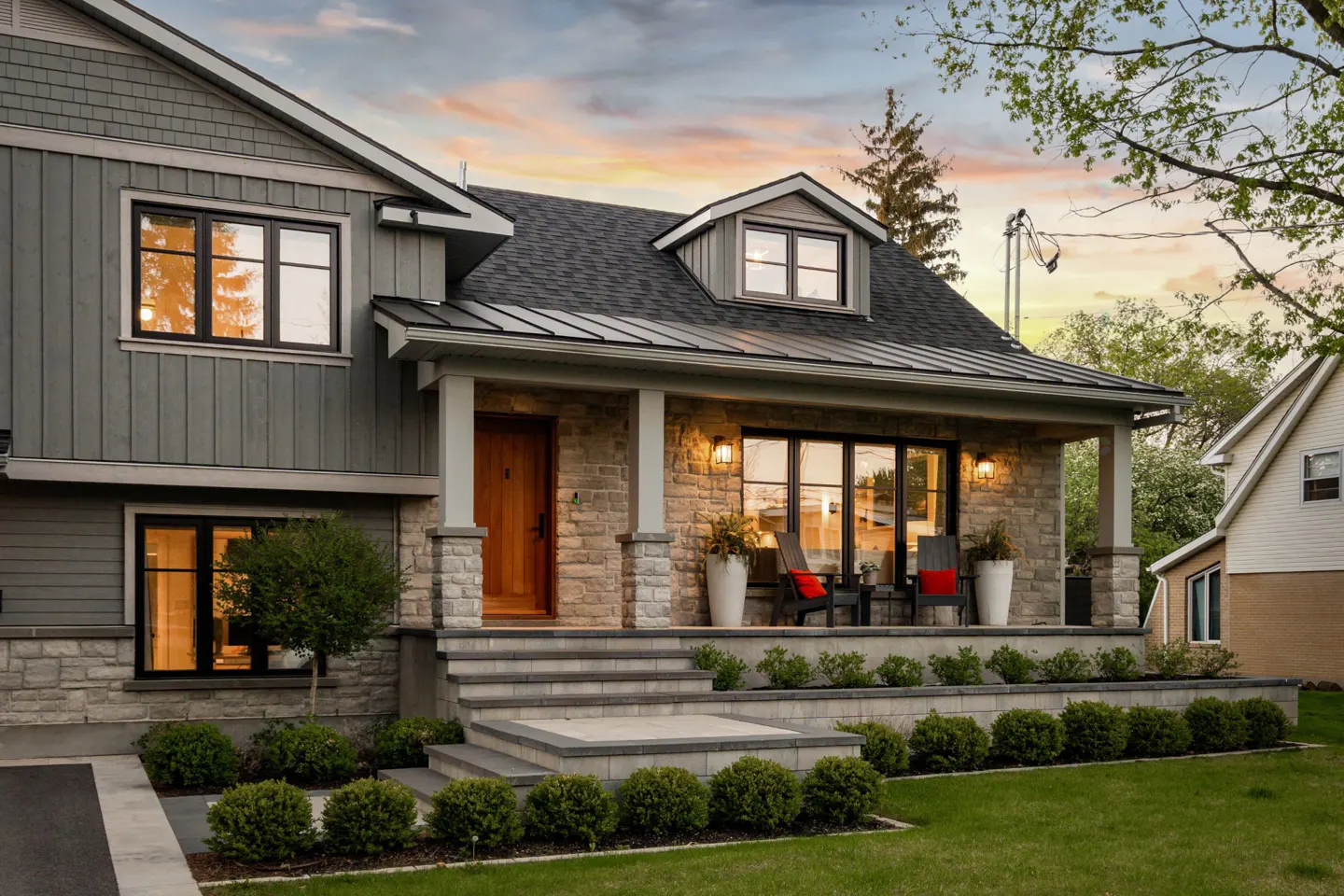 Two-story house with stone and gray siding, a covered porch with chairs, and manicured landscaping.