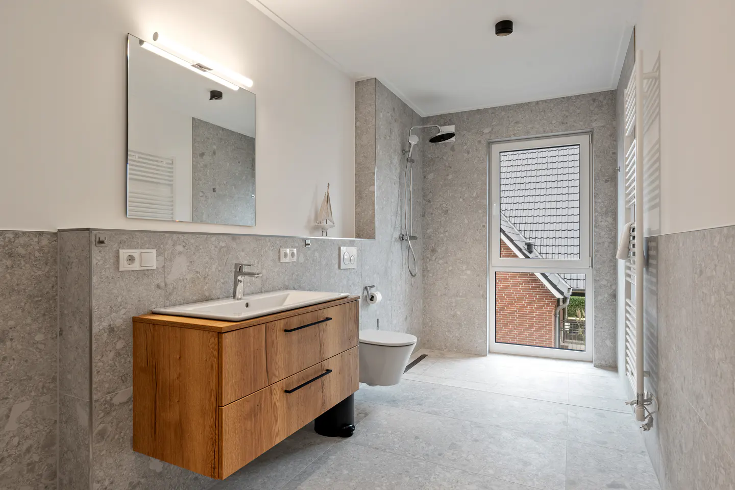 Modern bathroom with gray stone tiles, a wood vanity, white sink, toilet, shower, and a window with a view of a brick house.