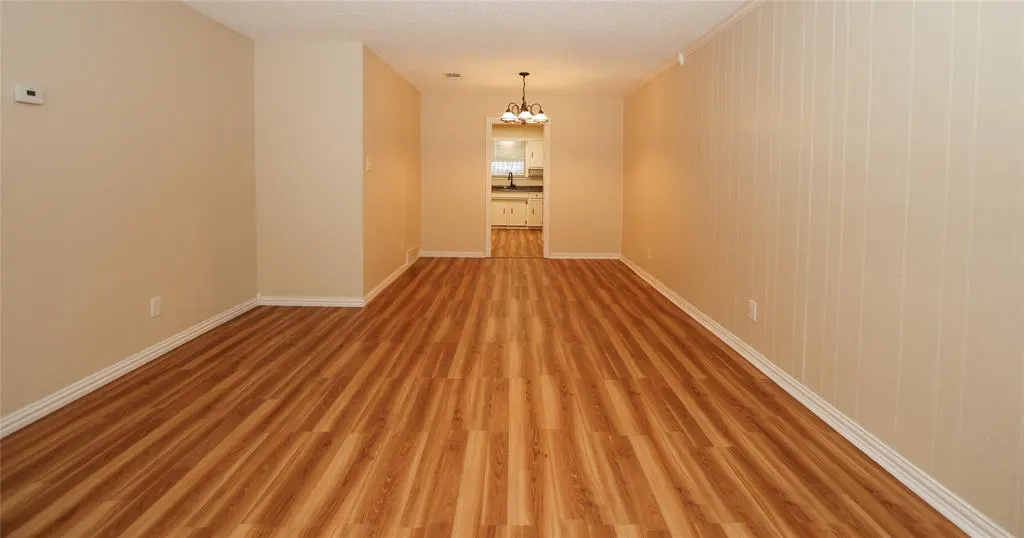 Empty room with wood-look flooring, beige walls, and white trim. A doorway leads to a kitchen with white cabinets.