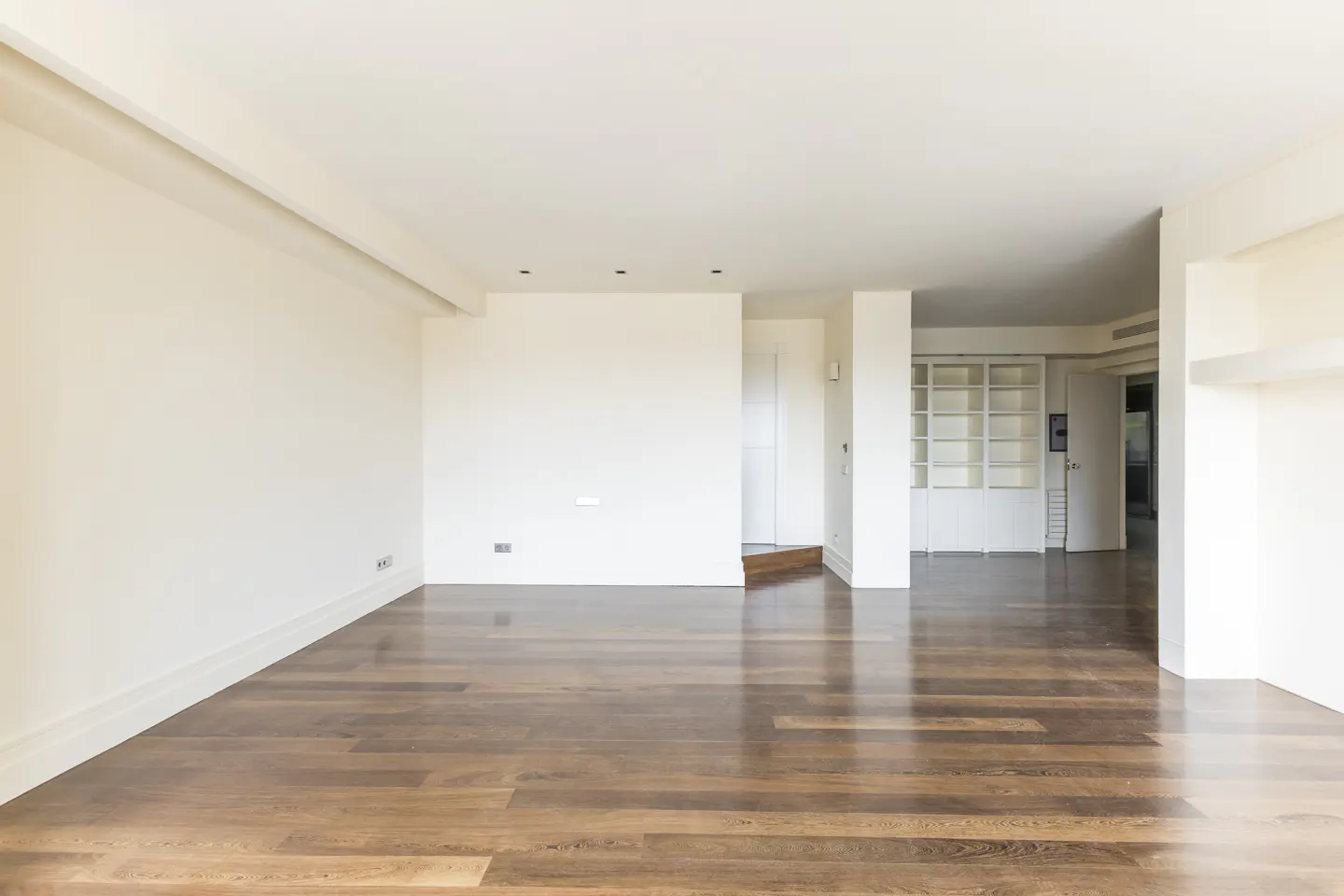 An empty room with hardwood floors and white walls. A built-in bookcase is visible in the background.