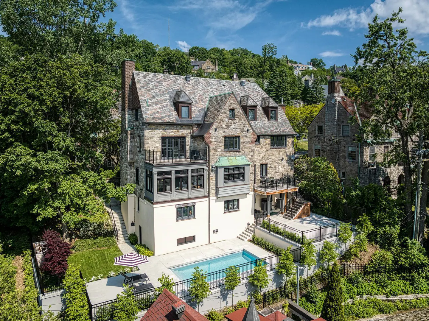 Aerial view of a stone house with a pool, patio, and balconies surrounded by trees.